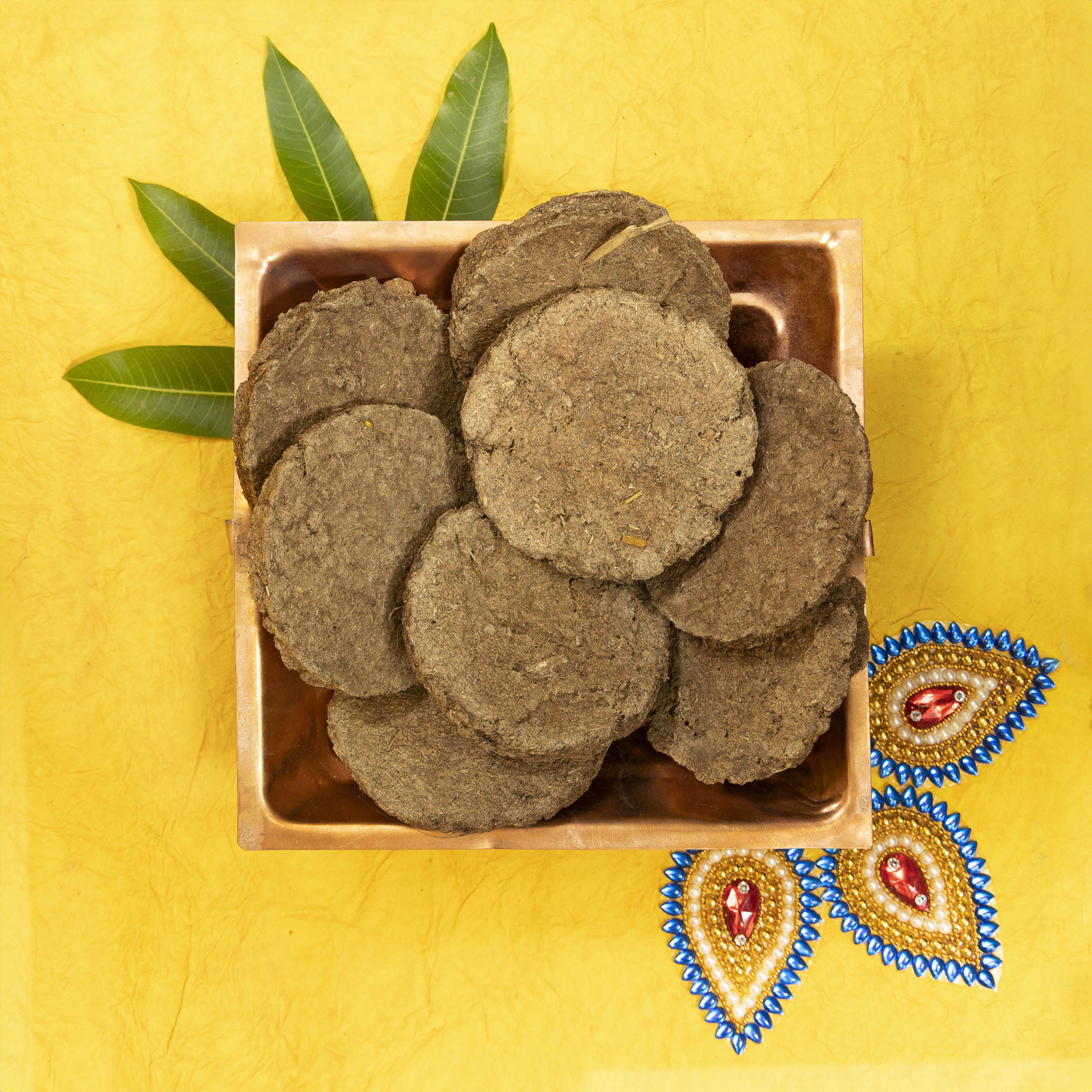 A top-down view of dark brown round herbal cakes, likely amla or amla powder cakes, arranged in a small square wooden dish on a textured yellow background with decorative green leaves and embroidered motifs.