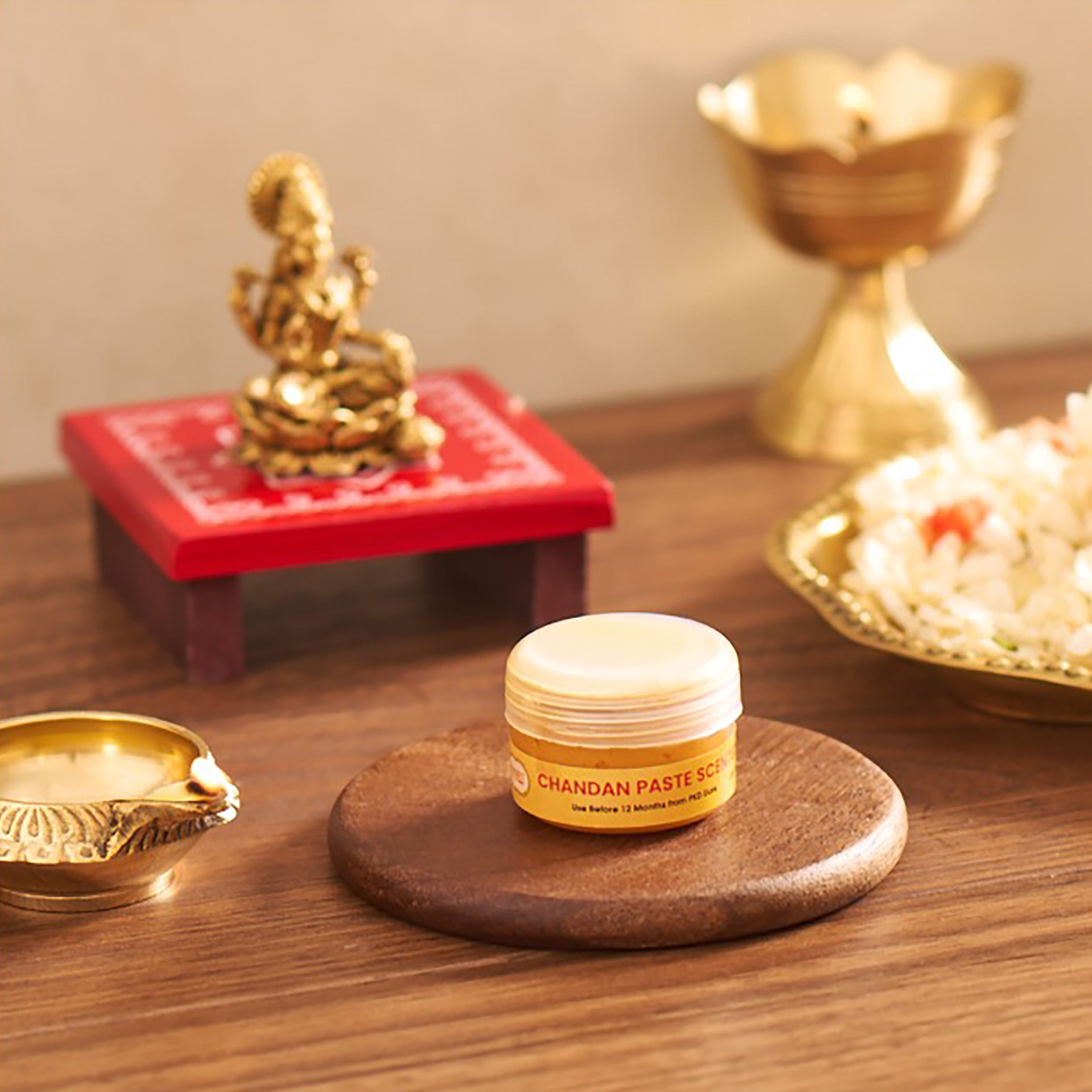 A small jar of Chandan Paste Kottam (holy ash) sits on a round wooden coaster on a wooden table, surrounded by traditional Indian decorative items including brass oil lamps, a red miniature stand with a gold statue, and a plate of rice.