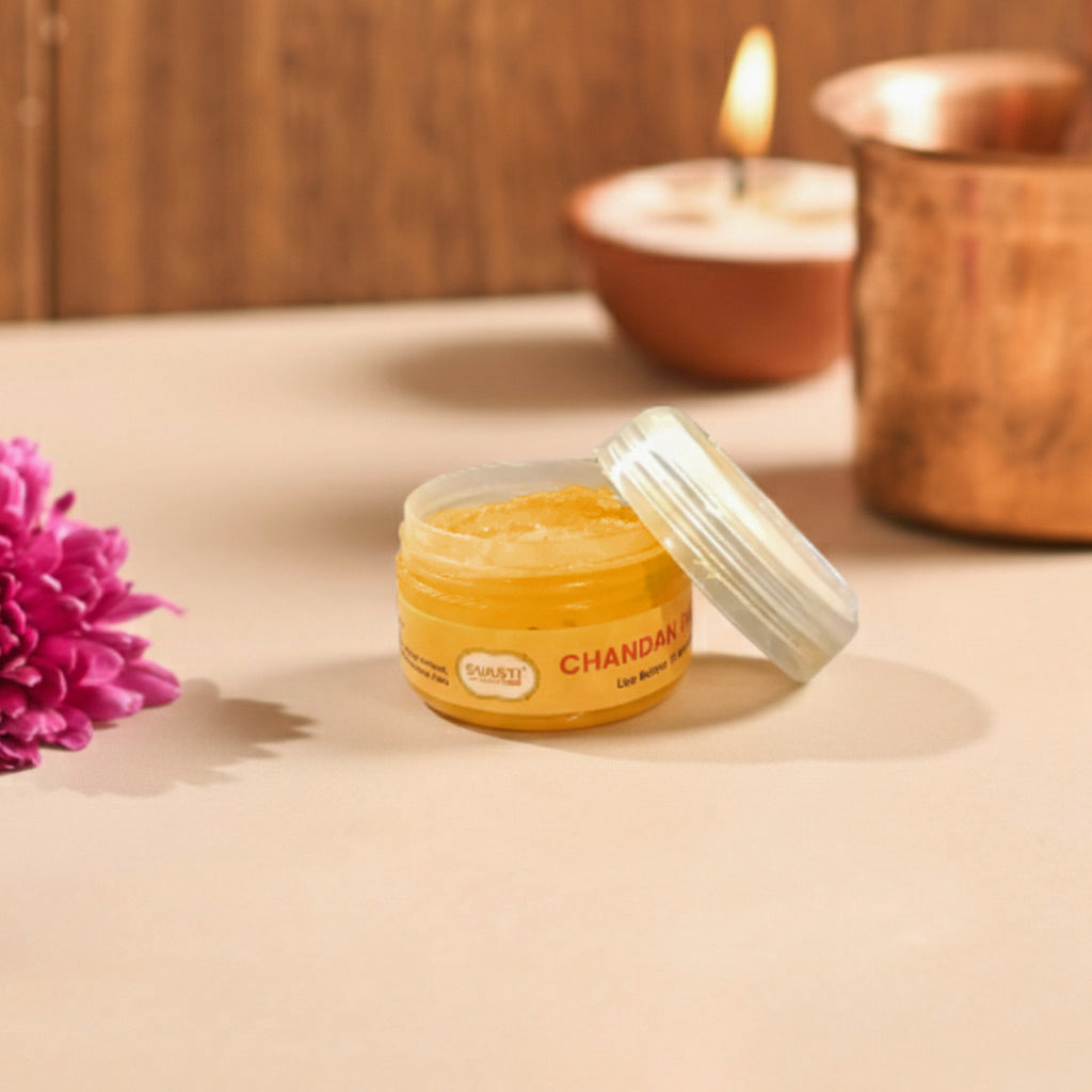 A jar of Chandali Shea Butter by the brand Parasu, shown with its lid off on a beige surface. The background features a warm, cozy setting with a lit candle in a terracotta bowl, a copper vessel, and a pink flower on the left.