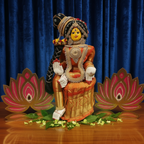 A decorative Hindu deity statue seated on a lotus throne, wearing an orange and gold sari with silver jewelry and a black-and-gold headdress, set against a bright turquoise background.