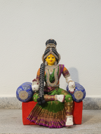 A colorful seated Hindu deity figurine, likely Lakshmi, wearing a green and purple sari with gold jewelry, sitting on a red block with blue cylindrical props, photographed against a plain off-white wall.