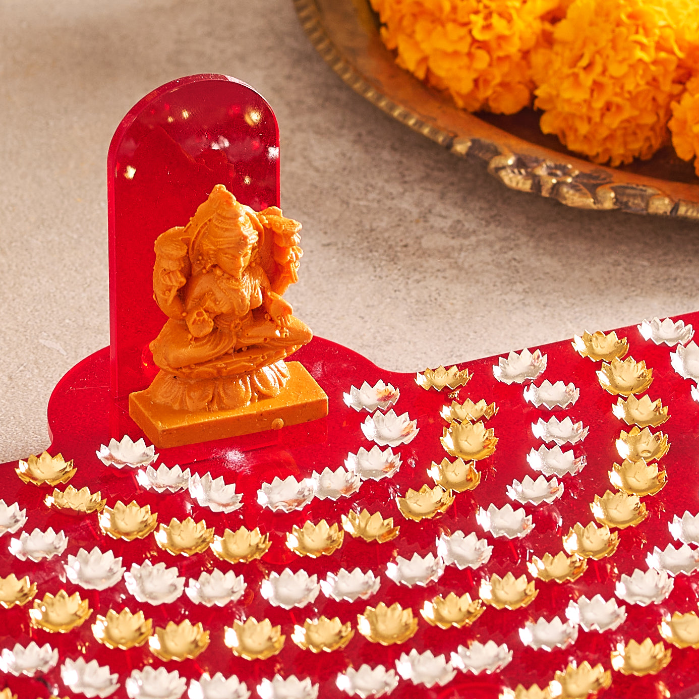 Small statue of a deity on a red platform with white and gold lotus flowers, placed on a textured surface.