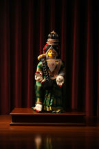A traditional Indian clay figurine of a seated goddess, likely Lakshmi, displayed on a wooden pedestal against a dark red curtain background.