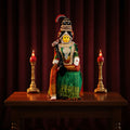A colorful idol of a female deity seated on a throne with a green and red design, holding a staff and a container, with gold accents and flowers in the background.