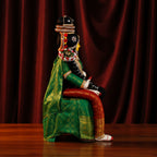 A traditional seated figurine or doll dressed in a green patterned robe with gold accents, red draped fabric on the legs, and a black decorated headpiece, posed against a dark red curtain backdrop.