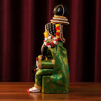 A traditional seated figurine or doll dressed in ornate green and gold patterned clothing, with a black hat, decorative beaded headpiece, red and yellow floral garlands, and white-and-red shoes, posed on a wooden surface against a dark red curtain background.