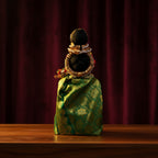 A traditional Indian doll with a black hair bun decorated with gold and white jewelry, wrapped in a green and gold patterned silk saree, standing on a wooden table against a dark red curtain background.