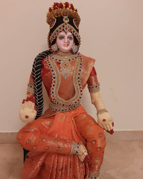 A seated decorative figurine or statue of a traditional Hindu deity, dressed in an orange and red sari with ornate gold jewelry, a black-and-gold crown, and painted skin, photographed against a plain beige wall.