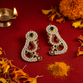 A pair of ornate gold-toned earrings embellished with clear stones, green and red gemstones, and white pearl-like beads, displayed on a red fabric background with orange flower petals, a small brass diya, and a pile of orange powder.