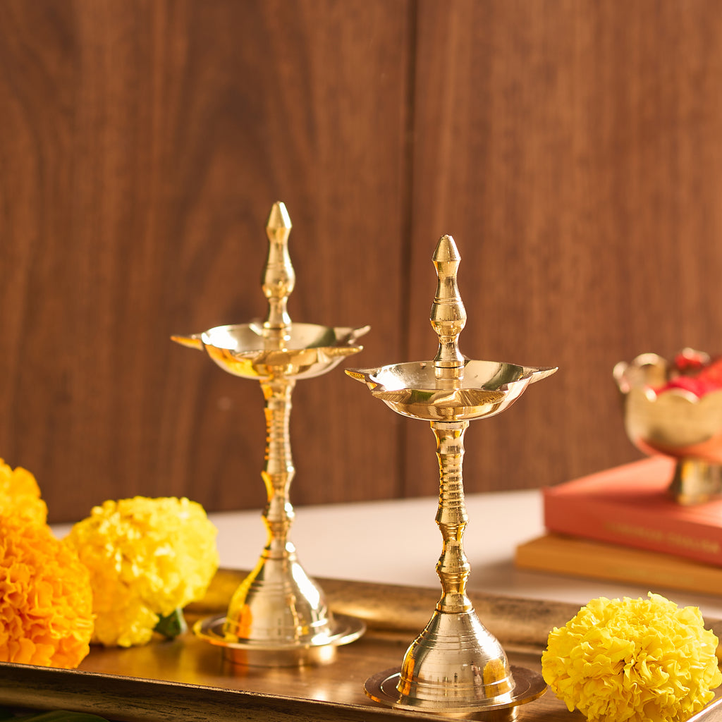 Two gold-colored oil lamps on a wooden surface with yellow flowers and books in the background.