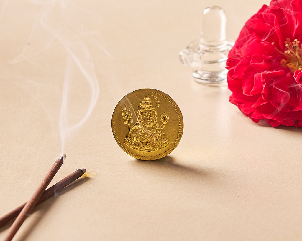 Gold coin with engravings on a beige surface with incense sticks, red flower, and glass container.