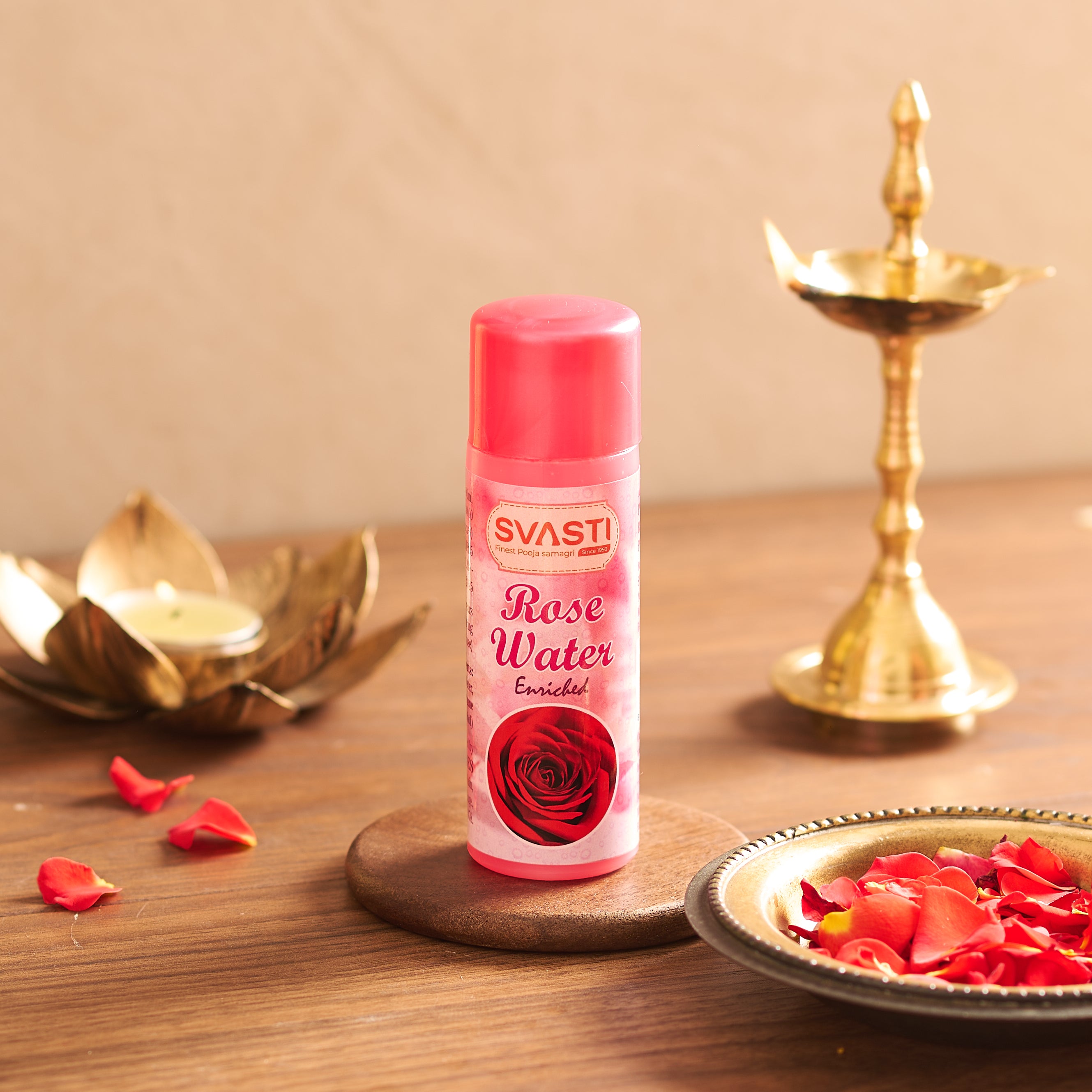 A slender cylindrical bottle of Svasti Rose Water featuring a bold magenta cap and a red rose illustration, resting vertically on a wooden table with blurred ritual garlands in the background.