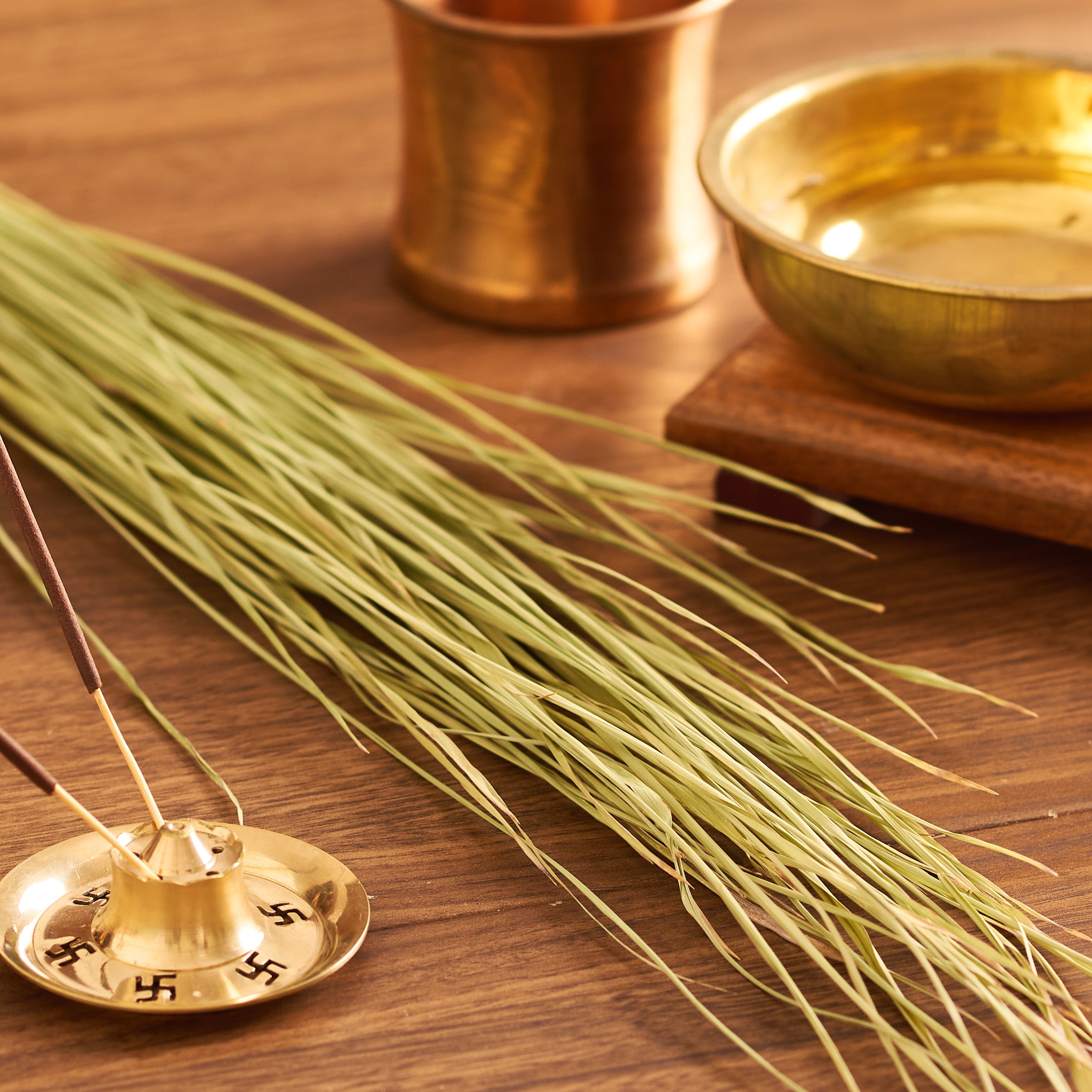 An incense stick with a holder, green grass, and brass cup and bowl on a wooden surface