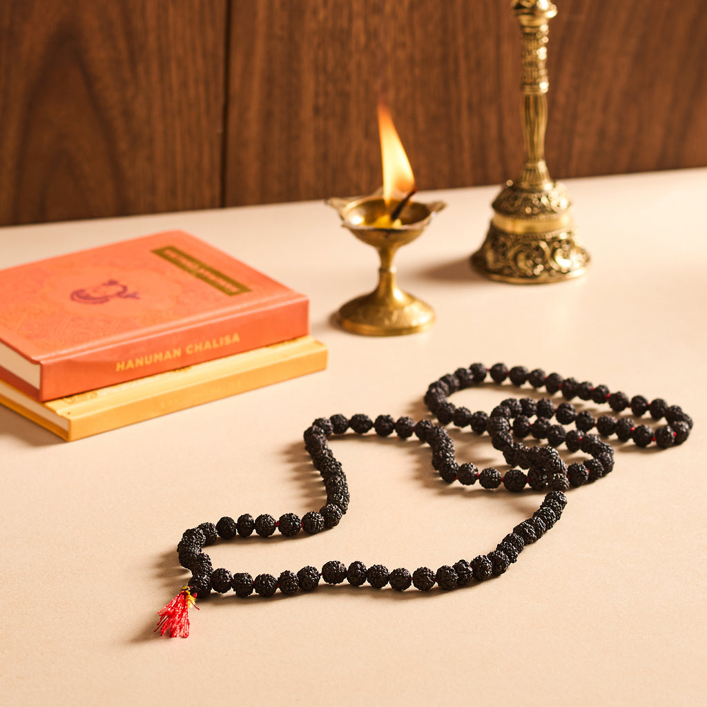 Black beaded necklace with a red tassel on a wooden surface with books and a lamp.
