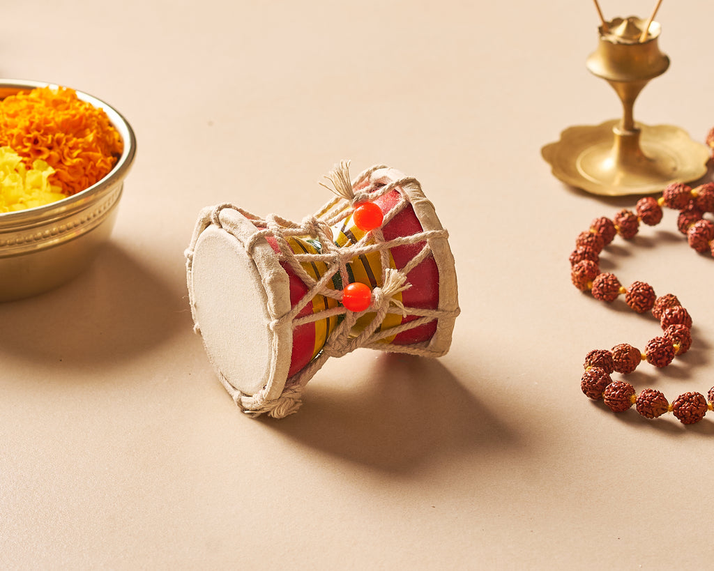 Small drum-like instrument with colorful beads on a beige surface, next to a bowl of yellow flowers and a brass lamp.