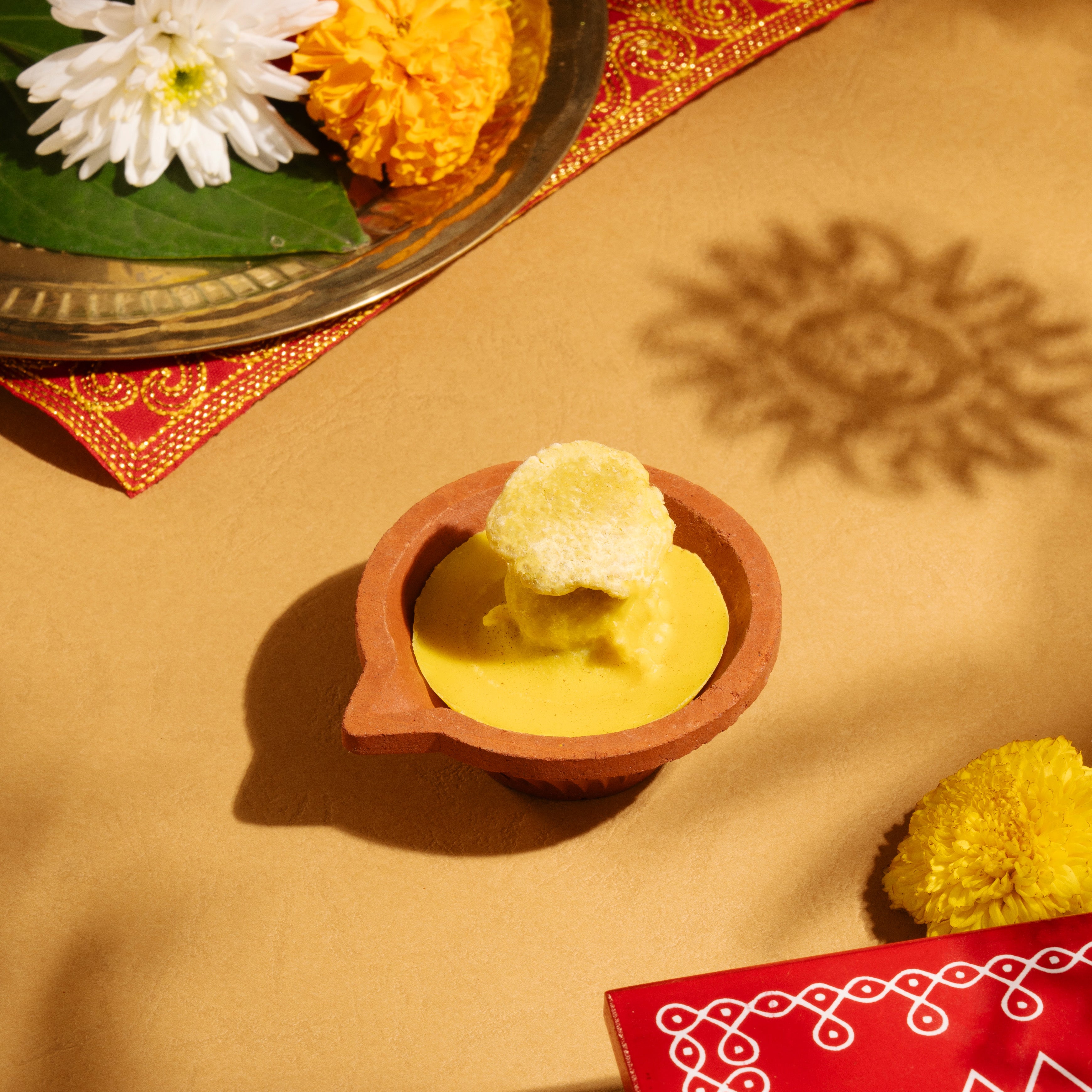 Front perspective view of the pre-filled ghee deepam clay diya resting on a surface next to its branded storage box and matchstick kit.