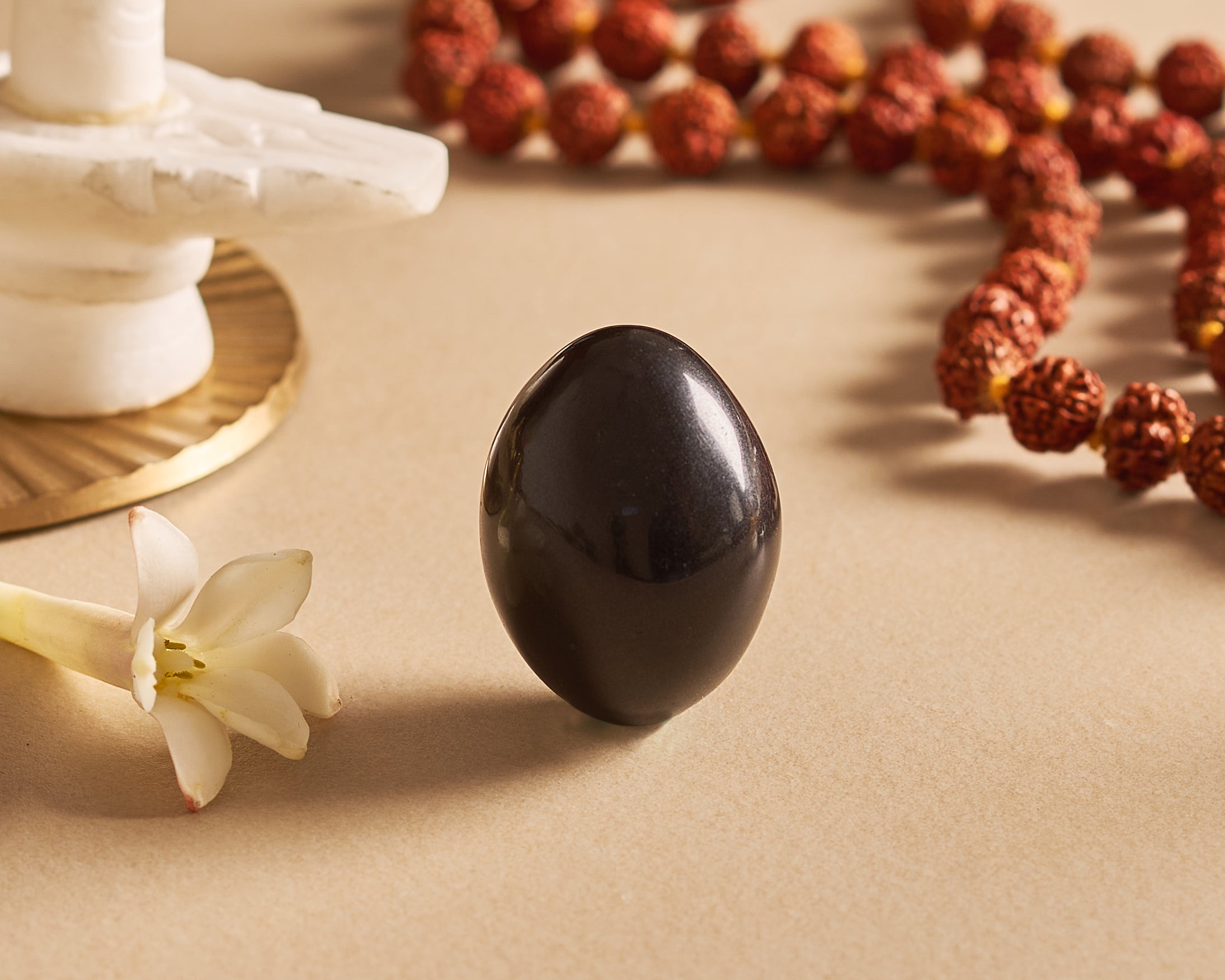 Black egg-shaped object on a beige surface with a white flower and red beads in the background