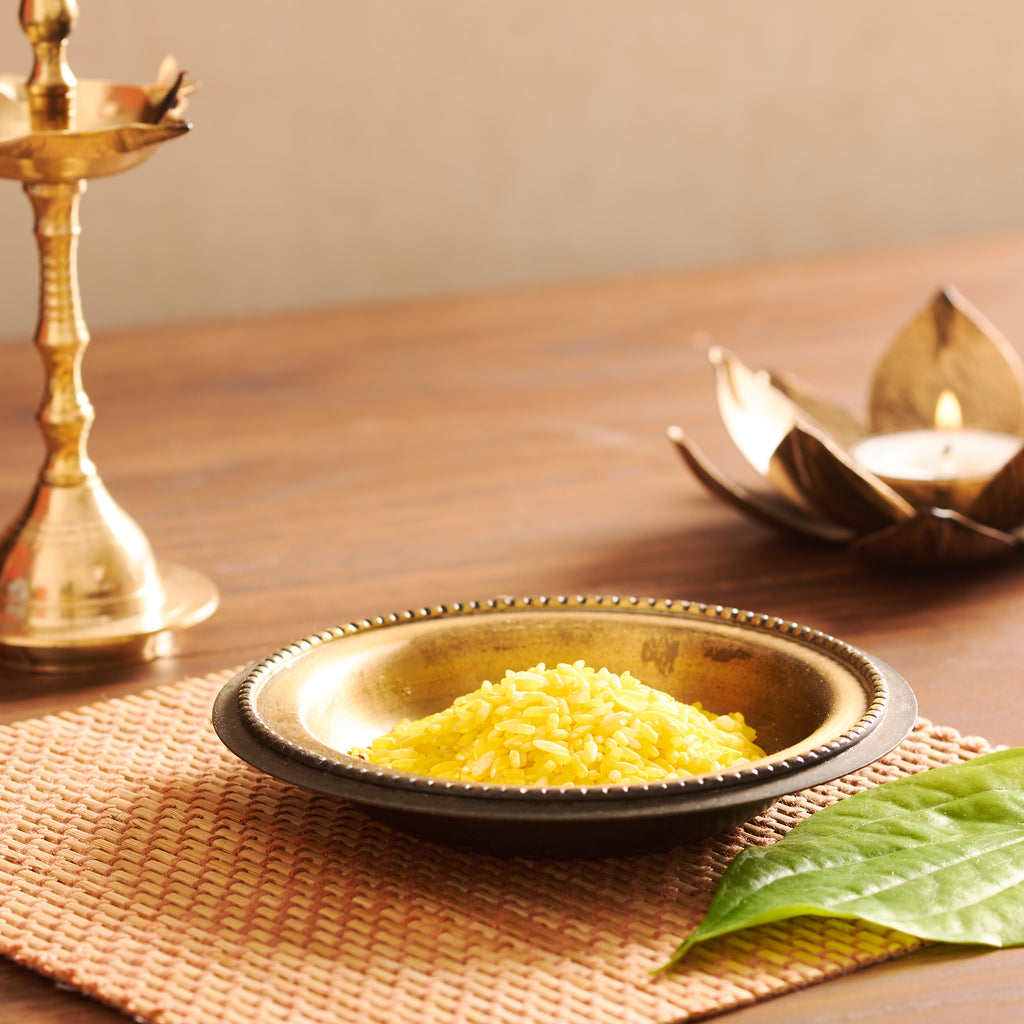 A neat, vibrant mound of lemon-yellow pooja rice resting in a traditional beaded brass plate on a textured mat, styled next to a fresh green betel leaf.