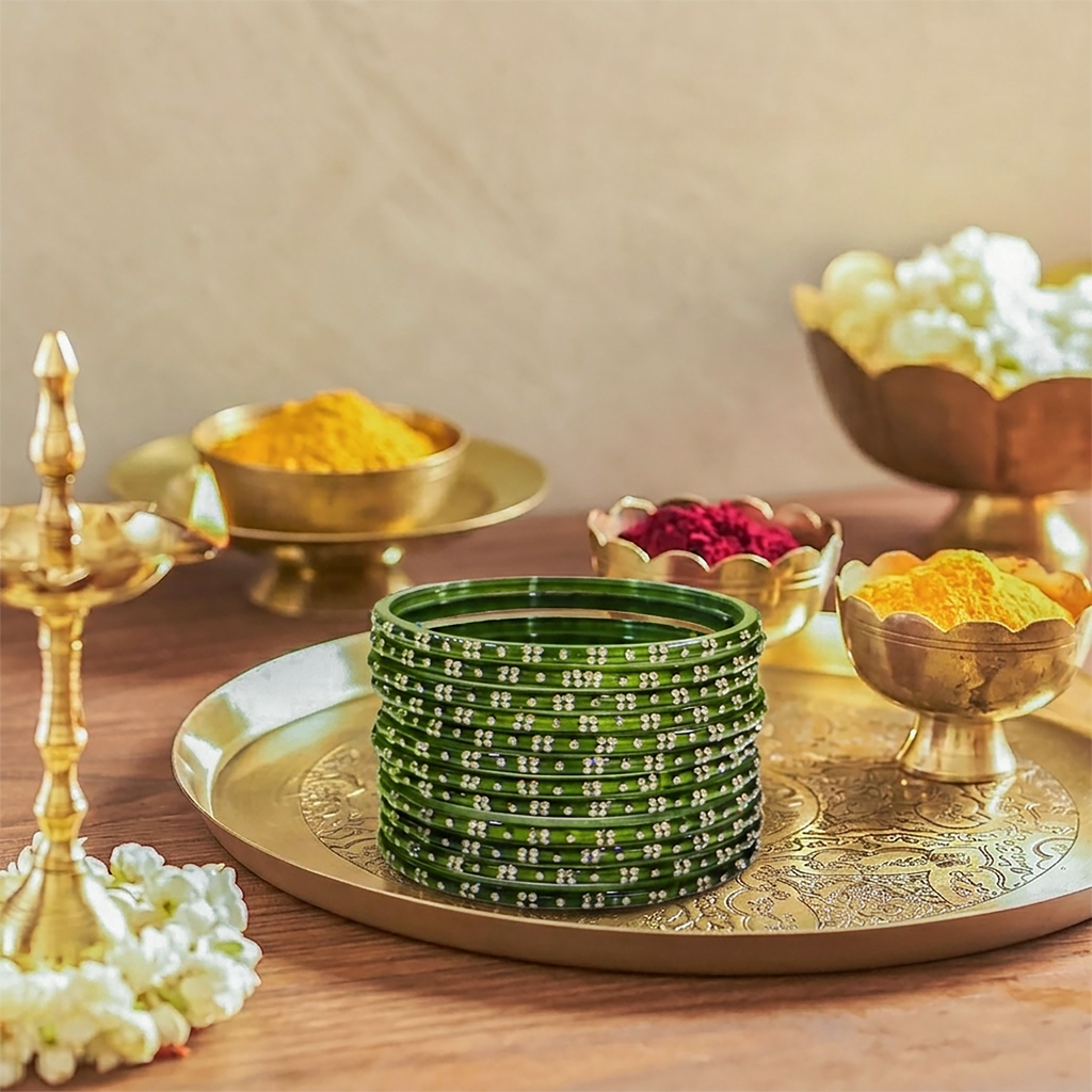 Stack of green bangles on a decorative tray with gold accents, surrounded by small bowls of colored powders and a lit lamp.