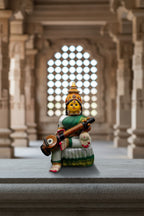 A colorful seated Hindu deity figurine with a yellow face, ornate gold and red crown, green sari, white skirt, and a stringed instrument, photographed against a dark backdrop with green artificial grass at the bottom.